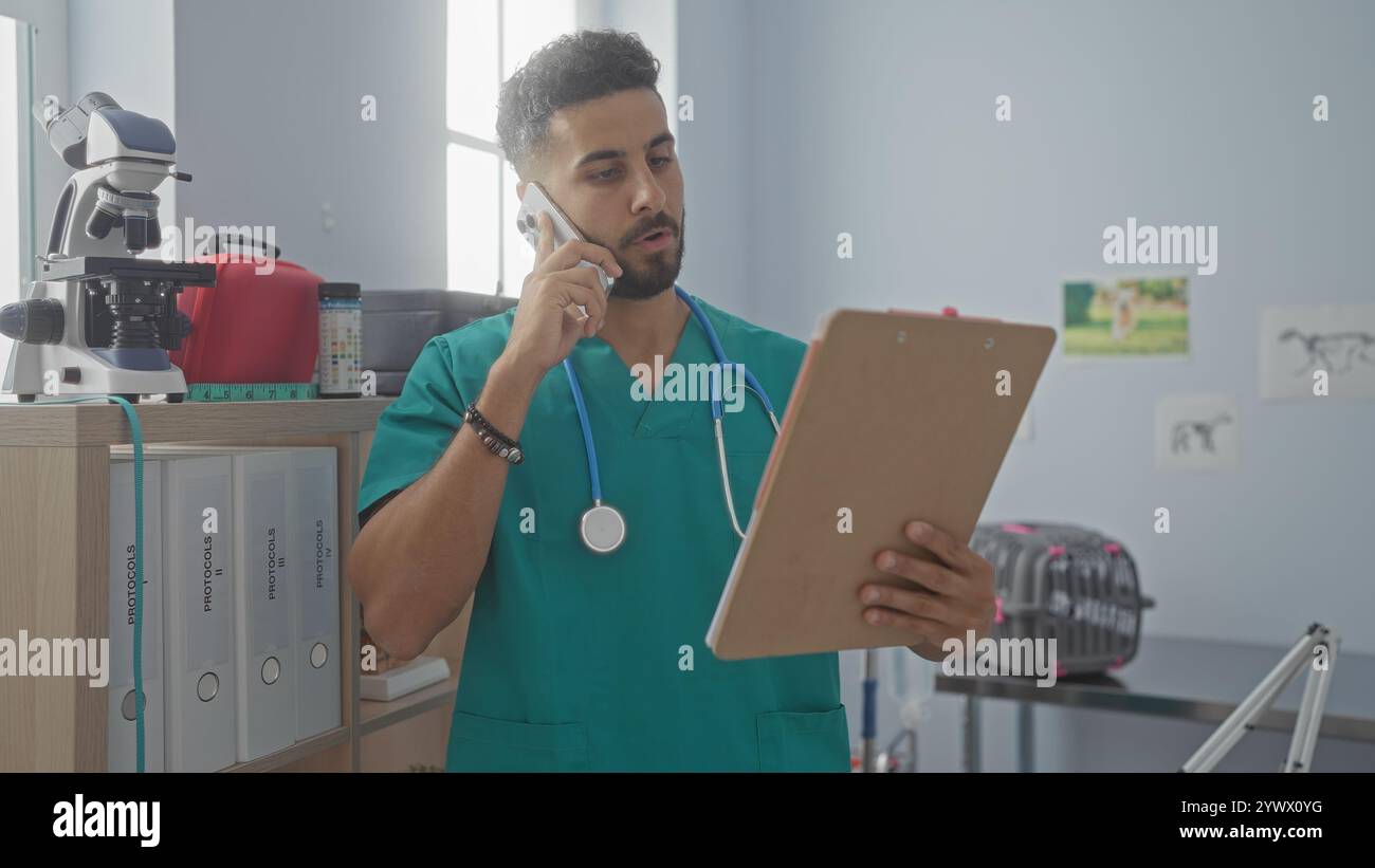 Un beau jeune homme hispanique dans une clinique vétérinaire examine une planche à pince tout en parlant au téléphone, portant des gommages verts et un stéthoscope autour du sien Banque D'Images