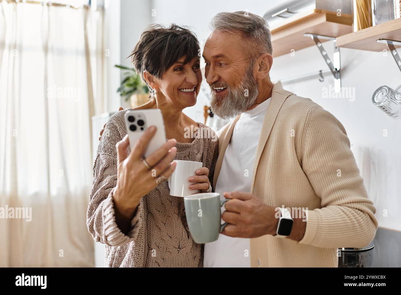 Couple senior partage un moment joyeux, tenant des tasses et capturant des souvenirs dans une cuisine ensoleillée. Banque D'Images