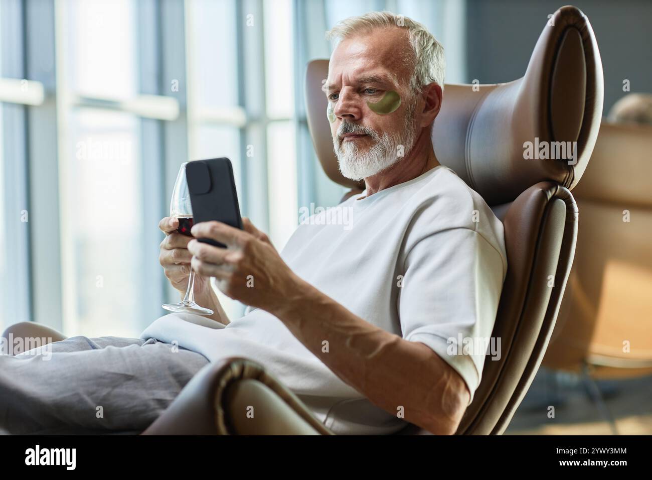 Plan moyen de l'homme d'affaires mature prenant soin de la peau du visage avec des cache-yeux, tout en envoyant des messages texte sur téléphone portable sur verre de vin rouge dans un fauteuil confortable à la maison Banque D'Images