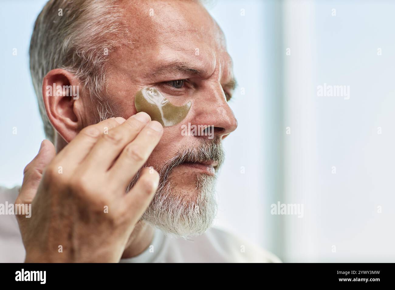 Portrait d'un homme âgé concentré appliquant soigneusement un patch hydrogel sous le masque de la doublure des yeux avec les doigts pendant la routine de soins de la peau à la maison, espace de copie Banque D'Images