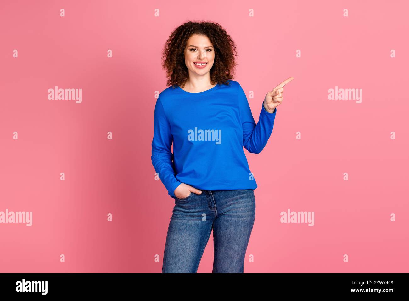 Jeune femme aux cheveux rouges en sweat-shirt bleu pointant avec un sourire sur un fond rose, favorisant des vibrations joyeuses Banque D'Images