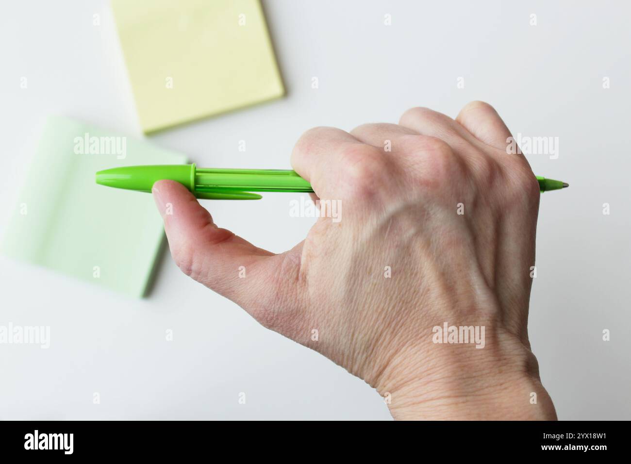 Stylo à bille vert tenu horizontalement par une main humaine caucasienne, notes collantes de couleur pastel en arrière-plan. Fournitures de bureau, activité d'écriture manuscrite. Banque D'Images