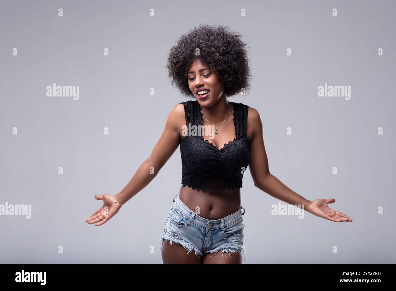 Jeune femme avec une coiffure afro haussa les épaules avec les bras ouverts et une expression confuse, regardant vers le bas et sur le côté contre un BA gris Banque D'Images