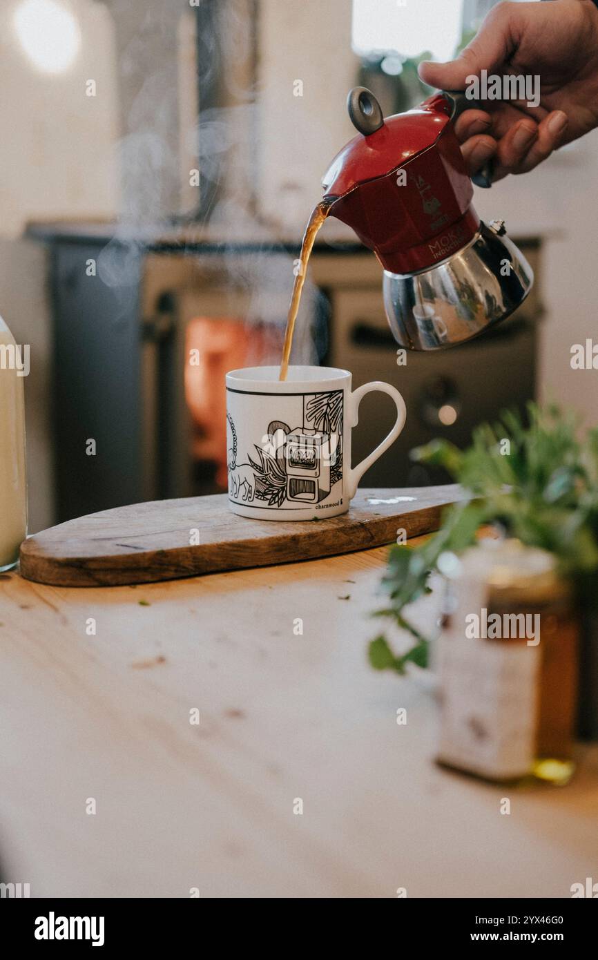 Une personne verse du café dans une tasse à partir d'une cafetière rouge. La cafetière est sur une table en bois à côté d'un pot de miel. La scène est confortable et tonique Banque D'Images