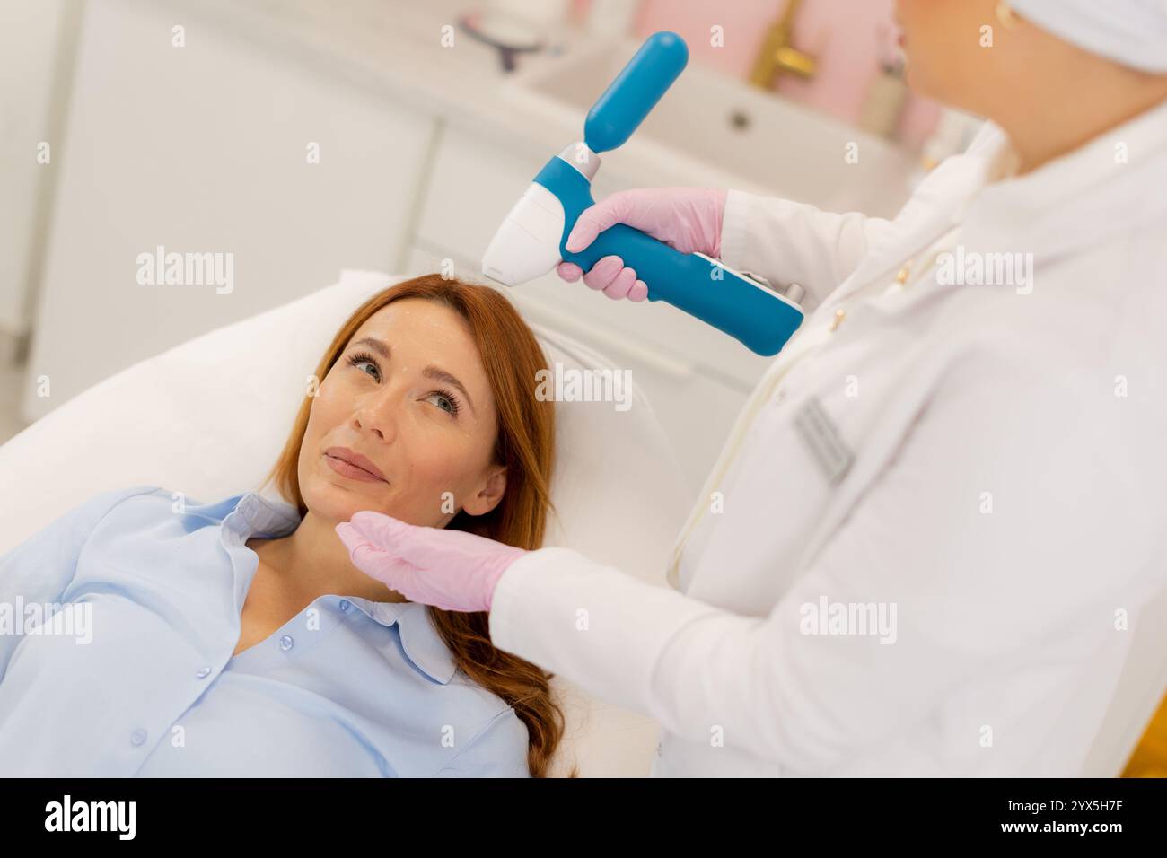 Le patient se détend sur un lit de traitement lorsqu'un technicien qualifié utilise un dispositif de refroidissement de la peau CO2 pour améliorer le confort pendant une procédure de rajeunissement du visage dans Banque D'Images