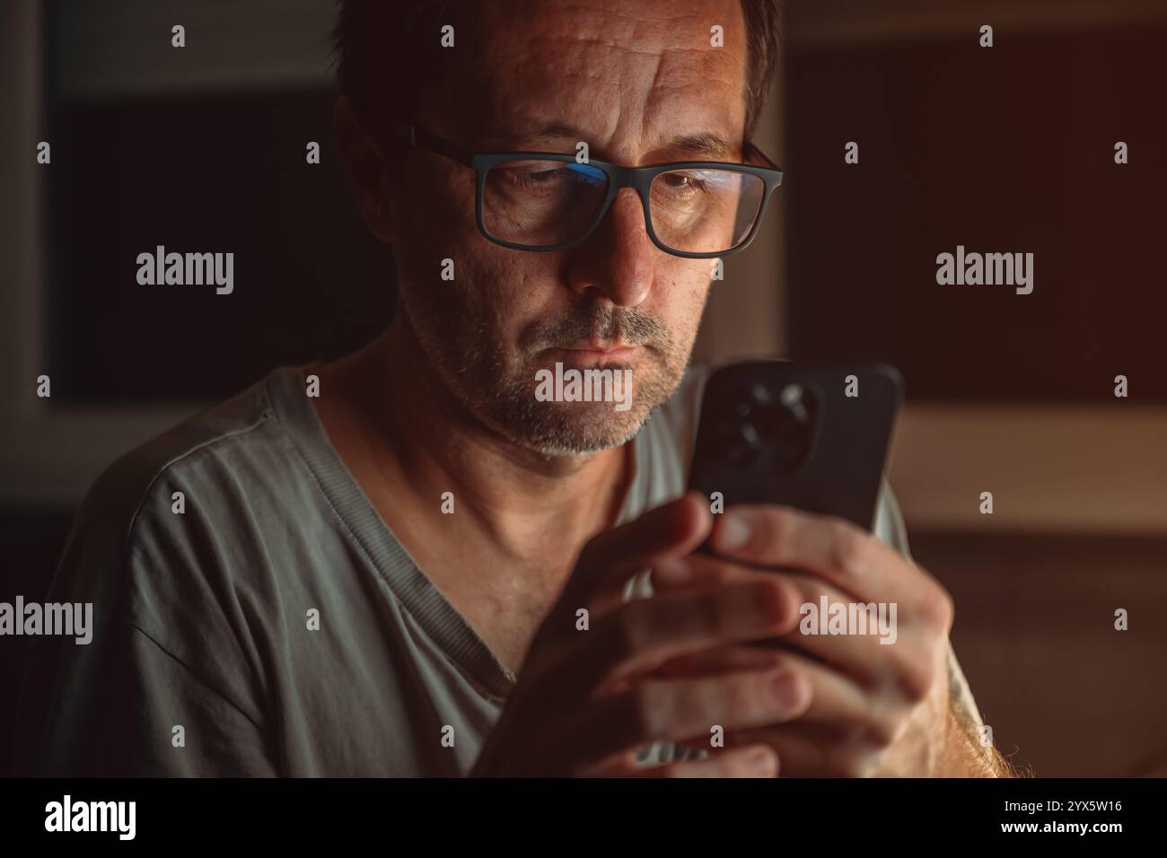 Adulte mature mâle dans le décontracté usé t-shirt répondant à un message texte sur son téléphone portable dans le confort de son salon tard dans la nuit, selecti Banque D'Images