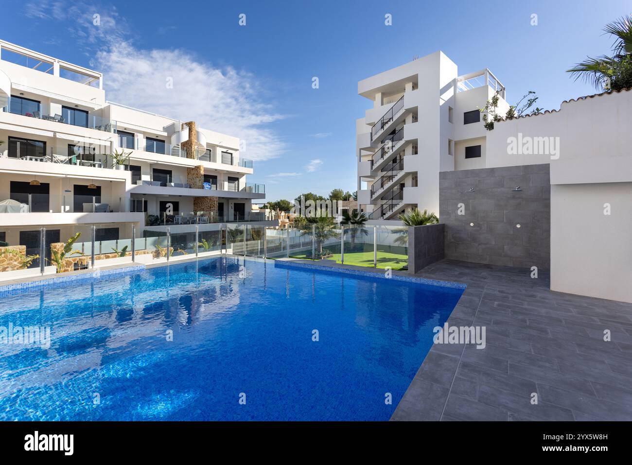 Complexe d'appartements élégant doté d'une grande piscine bleue, de balcons en verre, d'une architecture moderne et d'un cadre paysager sous un ciel dégagé. Banque D'Images