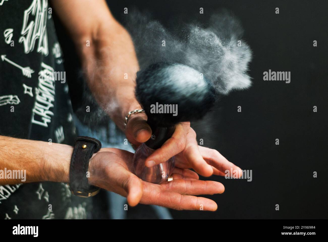 Les mains des hommes tiennent la brosse à raser à partir de laquelle le talc se disperse. Styliste masculin avec des outils de rasage sur un fond noir. Coiffeur au travail. Soin des cheveux et de la barbe. Banque D'Images