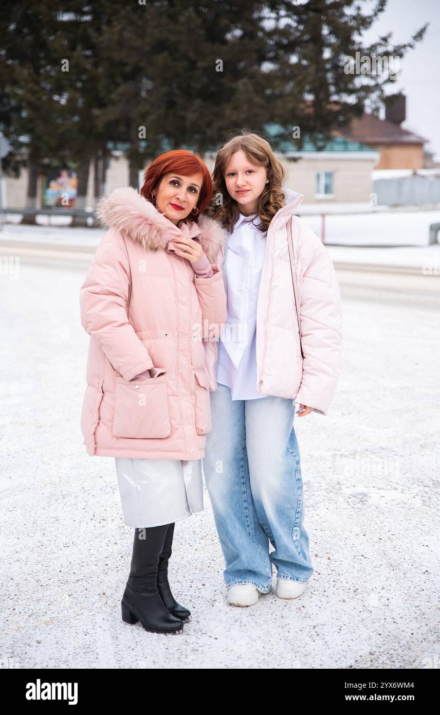 Mère célibataire russe avec sa fille adolescente debout dehors dans une ville enneigée. Les deux portent d'élégantes vestes d'hiver roses. Mode hivernale et famille co Banque D'Images