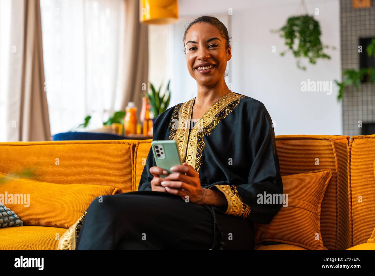 Heureuse belle jeune femme arabe portant traditionnel assis sur un canapé à la maison en utilisant un téléphone intelligent pour magasiner en ligne Banque D'Images