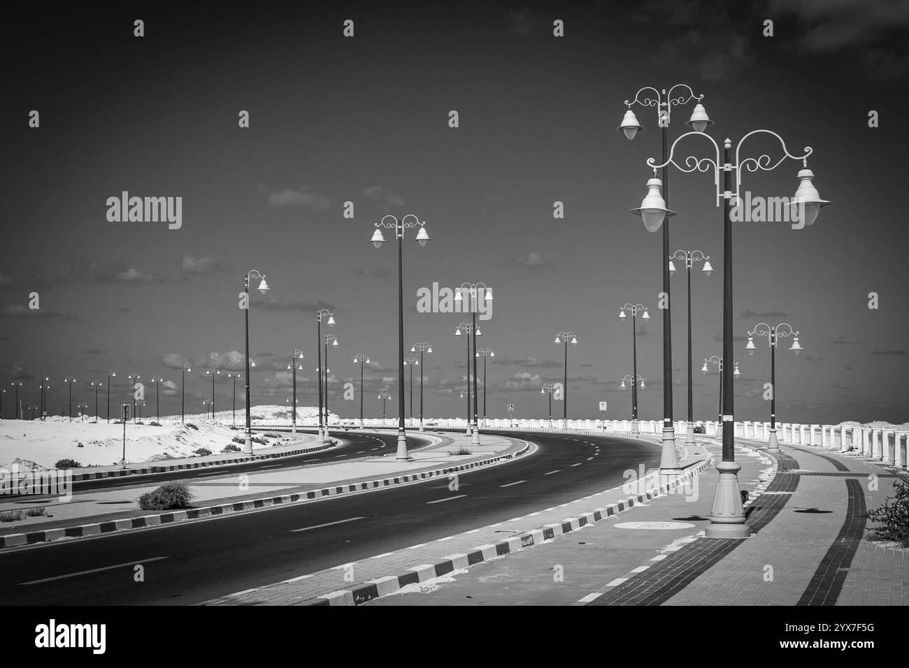 Une image captivante en noir et blanc de la paisible promenade côtière de Mersa Matruh, avec d'élégants lampadaires et une route courbe contre le ciel. Banque D'Images