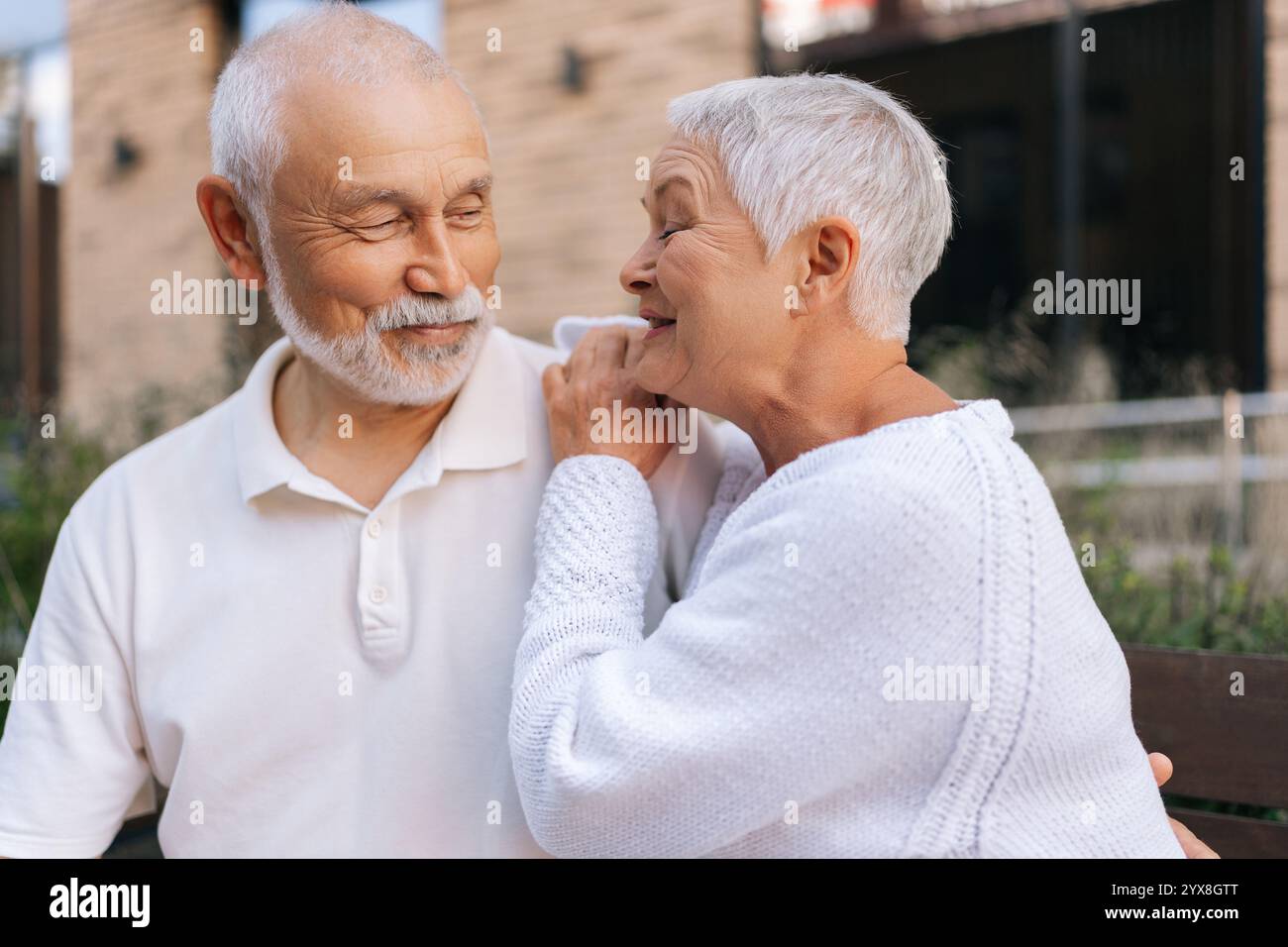 Portrait de belle femme âgée aux cheveux gris touchant doucement l'épaule partenaire partageant un moment chaud et aimant assis sur le banc de la ville le jour d'été. Banque D'Images