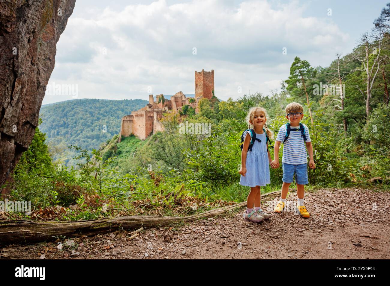 Devant une forteresse historique Saint-Ulrich et des collines verdoyantes, deux enfants heureux avec des sacs à dos posent sur un chemin en Europe, région Alsace, France Banque D'Images