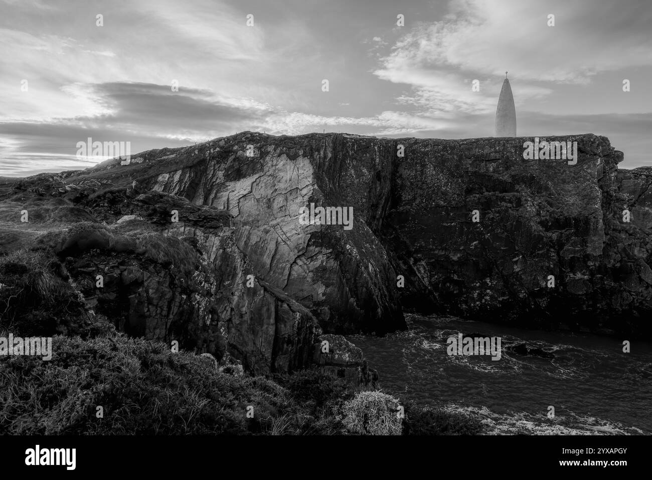 L'image montre une pente de roches gris foncé et d'herbe. Une tour blanche, mince et lointaine est visible. Banque D'Images