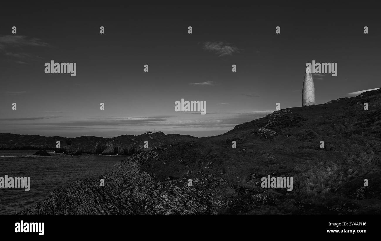 Un marqueur blanc, haut et mince se dresse sur une falaise herbeuse. Des roches sombres et accidentées forment la falaise. La mer s'étend jusqu'à un littoral lointain. Banque D'Images