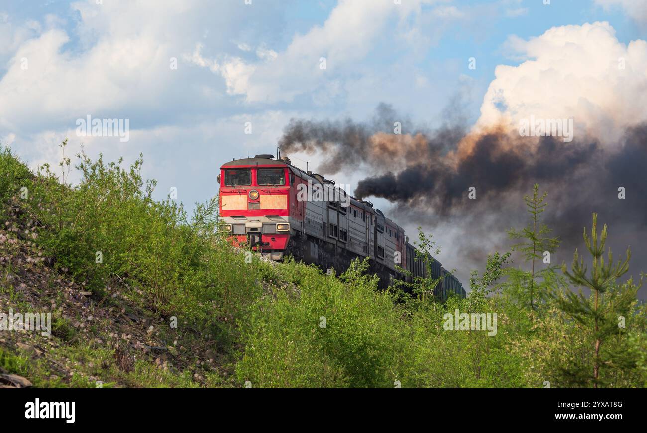 Un train de marchandises rouge se déplace le long d'un remblai contre le ciel, dégageant des panaches de fumée Banque D'Images Un train de marchandises rouge se déplace le long d'un remblai contre le ciel, dégageant des panaches de fumée Banque D'Images