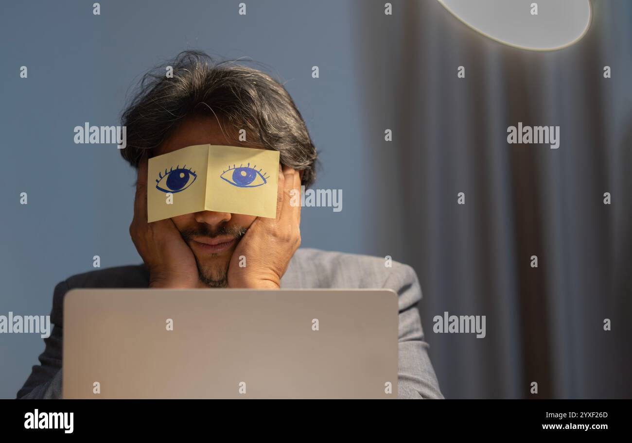 Les employés masculins asiatiques paresseux ont des autocollants avec des yeux peints se sentent submergés par le travail informatique au bureau la nuit. Banque D'Images
