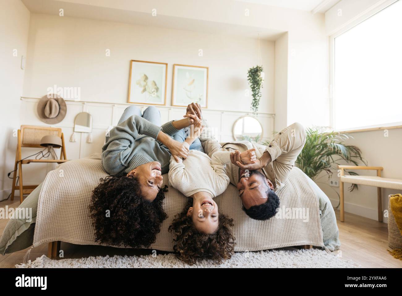 Une famille joyeuse passe du temps de qualité ensemble à la maison tout en jouant à l'envers sur un lit. Les parents et l'enfant partagent le rire dans une chambre confortable. Banque D'Images