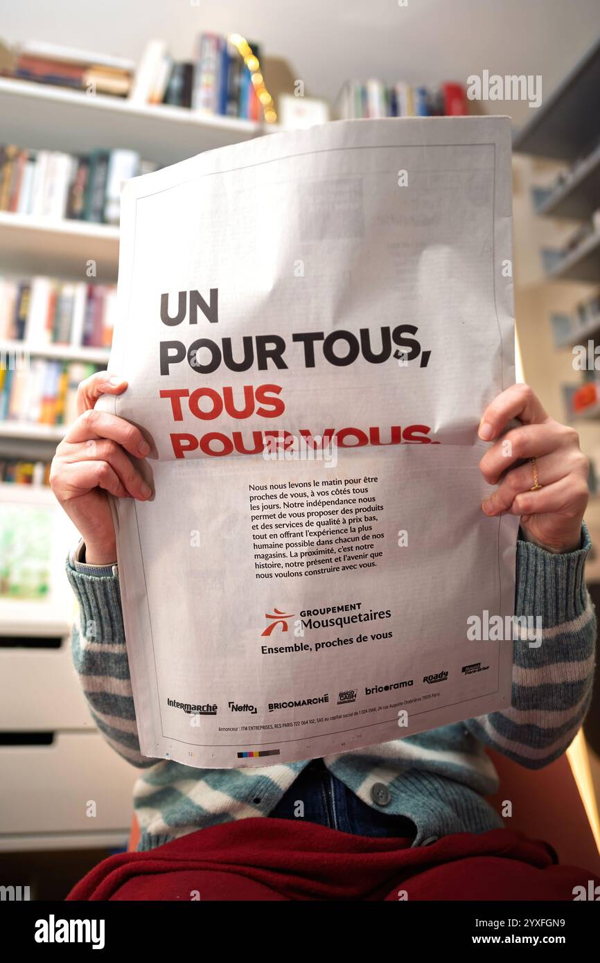 Personne tenant un journal avec une publicité pour groupement Mousquetaires avec le slogan « un pour tous, tous pour vous » dans un cadre intérieur confortable Banque D'Images