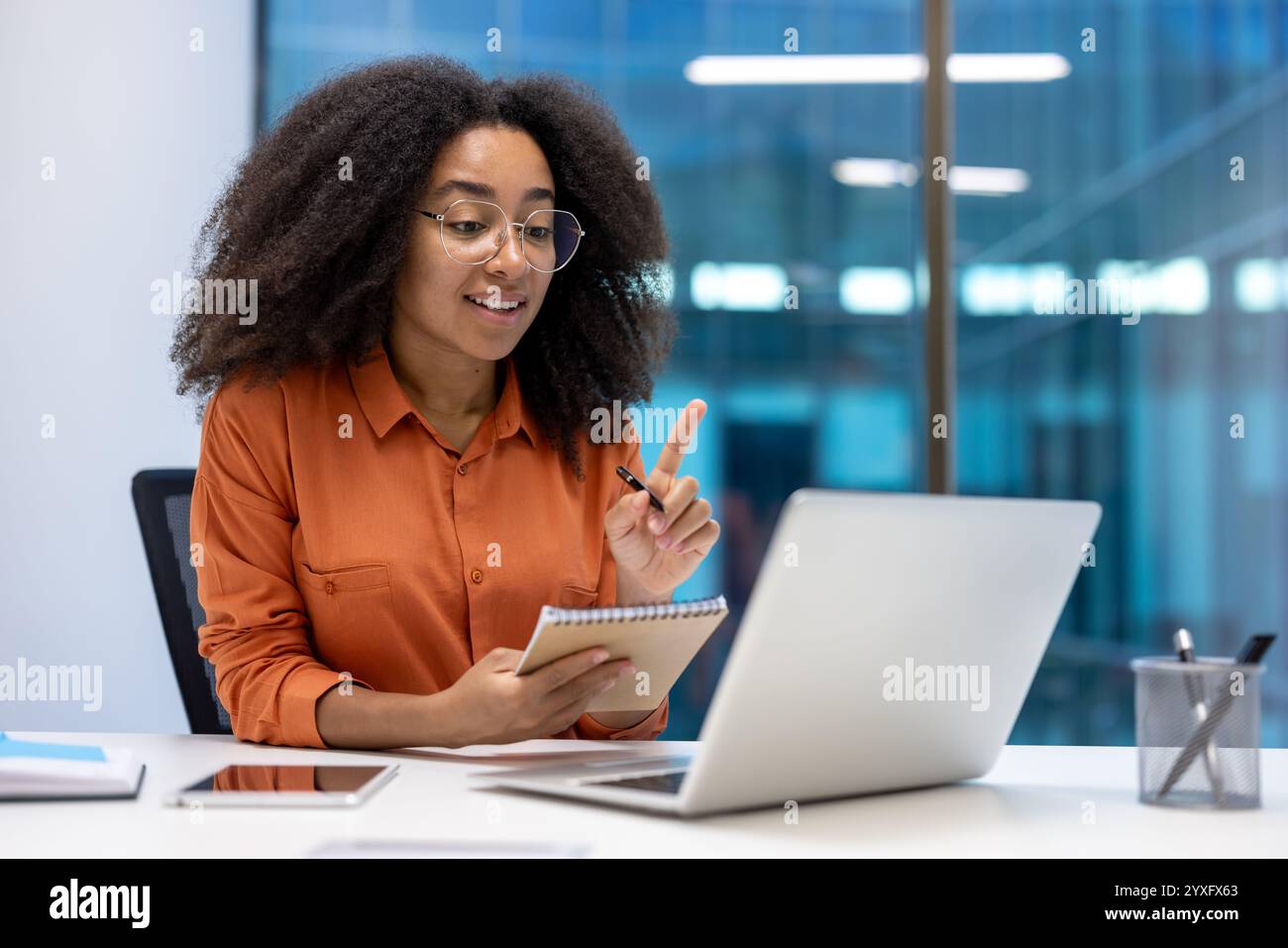 Un professionnel participe à une réunion virtuelle à l'aide de son ordinateur portable et note des notes dans son ordinateur portable. Elle est dans un cadre de bureau moderne avec de grandes fenêtres en verre Banque D'Images