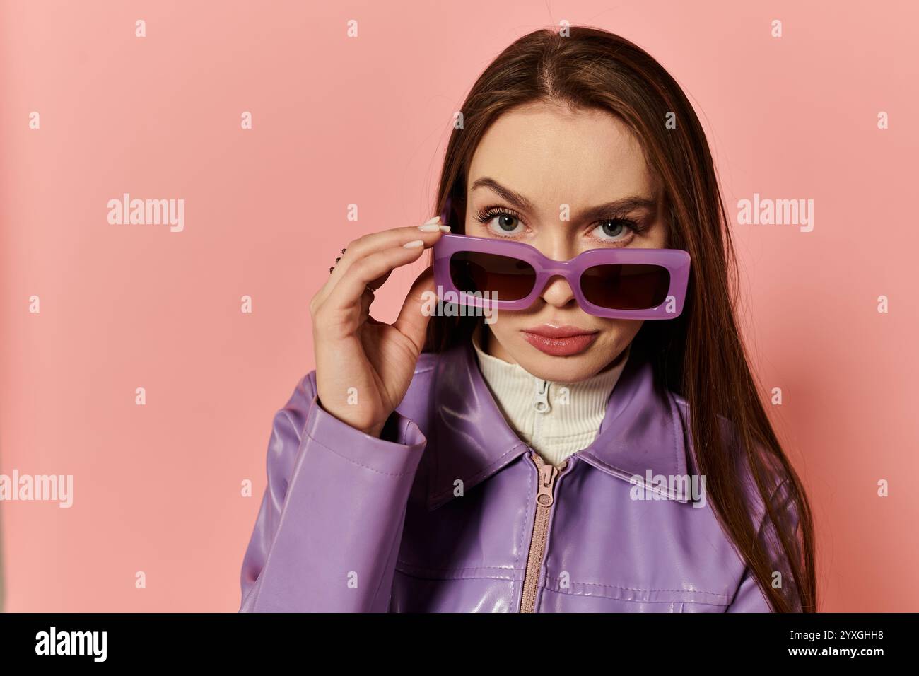 Une femme élégante ajuste de manière ludique ses lunettes de soleil surdimensionnées sur un fond rose. Banque D'Images