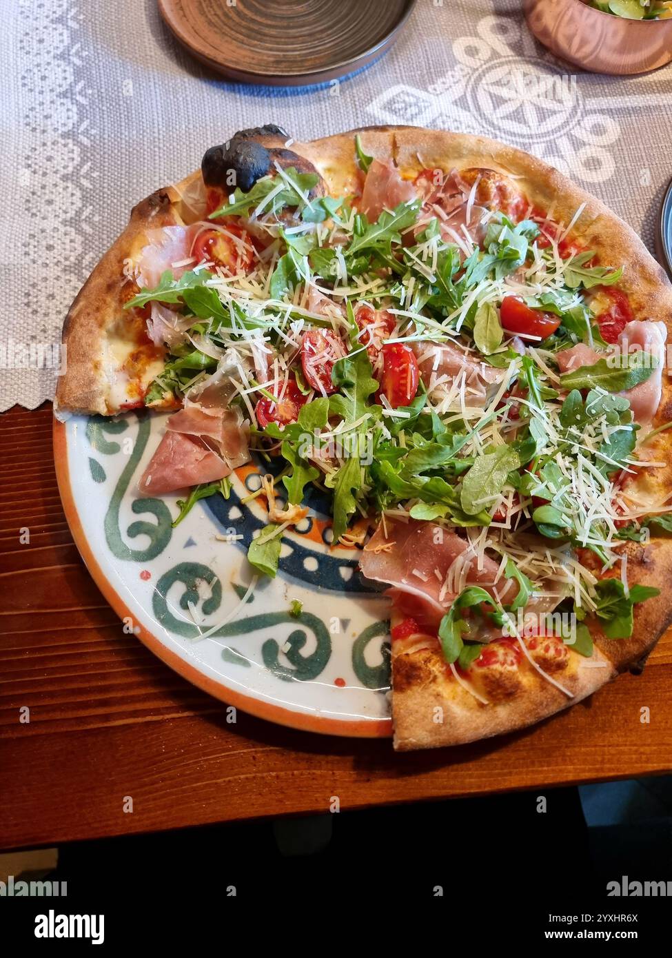 Pizza italienne premium garnie de roquette, prosciutto, tomates cerises et parmesan sur une assiette décorative, partiellement mangée, vue à la première personne, Banque D'Images