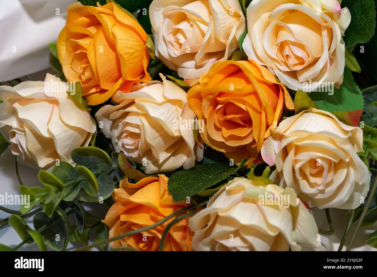 Un bouquet de roses artificielles présente des nuances d'orange et de crème. Décorées de feuillage vert, ces fleurs réalistes apportent une touche d'élégance à un Banque D'Images