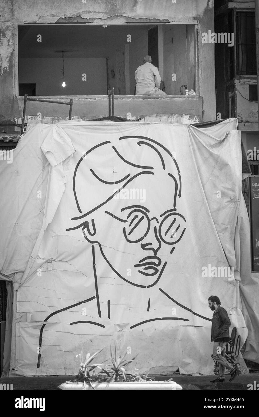 Une grande murale en noir et blanc représentant une femme portant des lunettes de soleil se drape sur un chantier de construction à Alexandrie, en Égypte, avec un passant. Banque D'Images