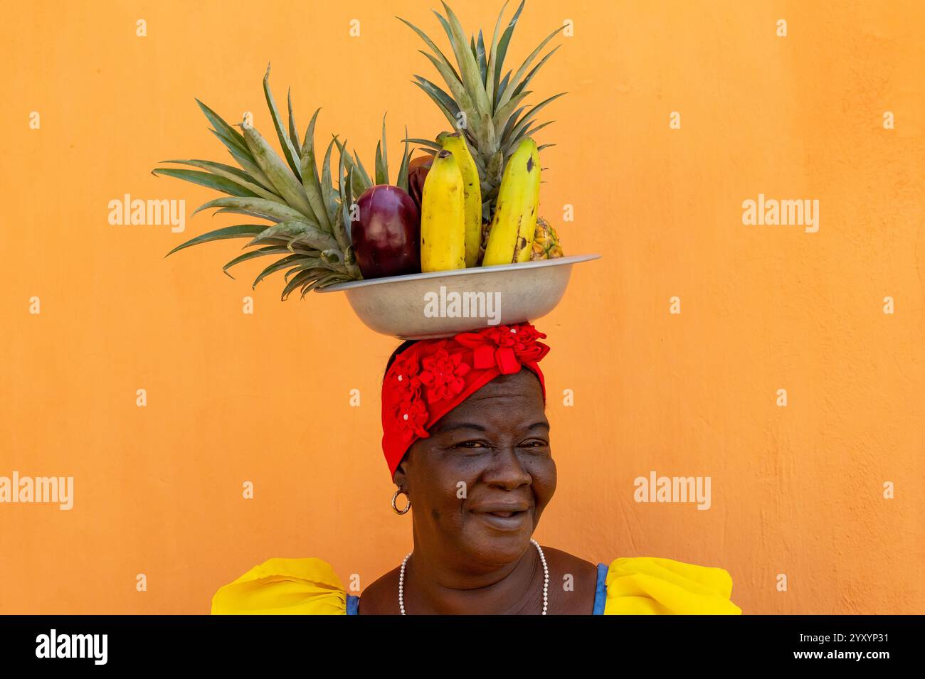 Femme dans Une robe colombienne colorée équilibrant Un bol de fruits tropicaux contre un mur orange Banque D'Images