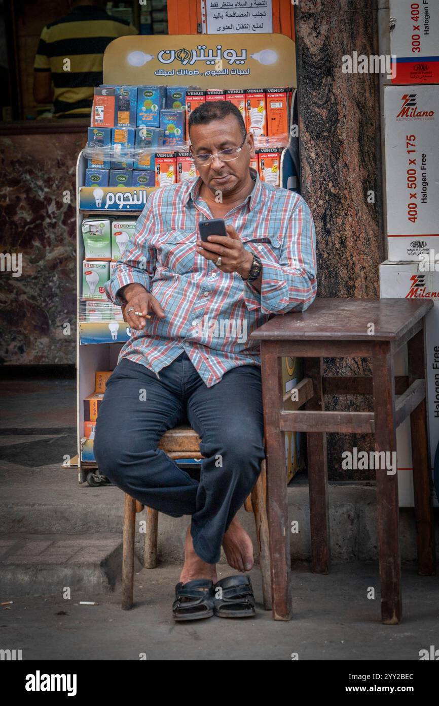 Un homme à Alexandrie, en Égypte, est assis devant un magasin vendant des ampoules et des fournitures électriques, utilisant son téléphone. Scène de rue détendue de la vie quotidienne. Banque D'Images