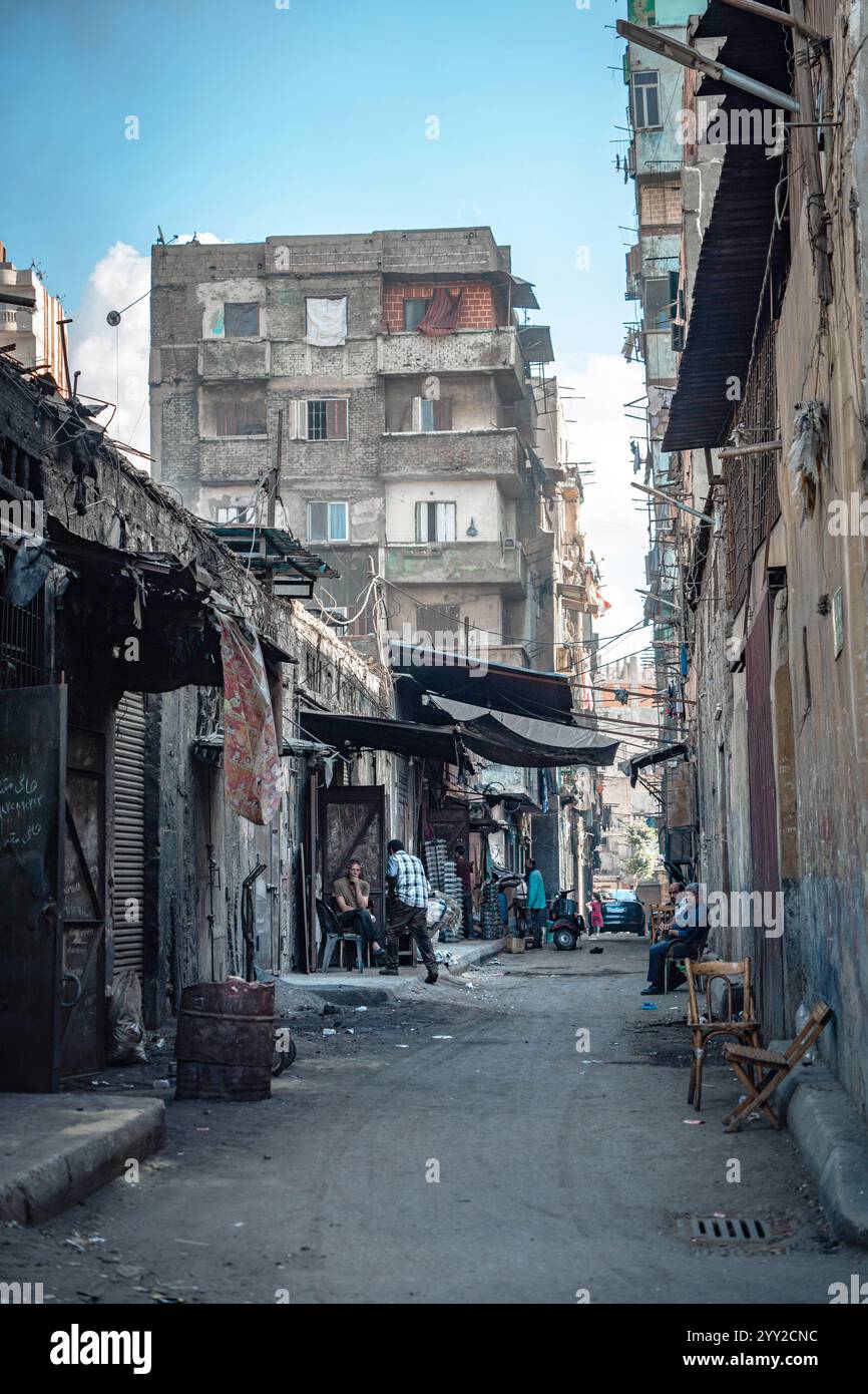 Ruelle étroite à Alexandrie, en Égypte, bordée de vieux bâtiments, de boutiques de rue et de résidents se détendant sur des chaises, capturant la vie urbaine égyptienne. Banque D'Images