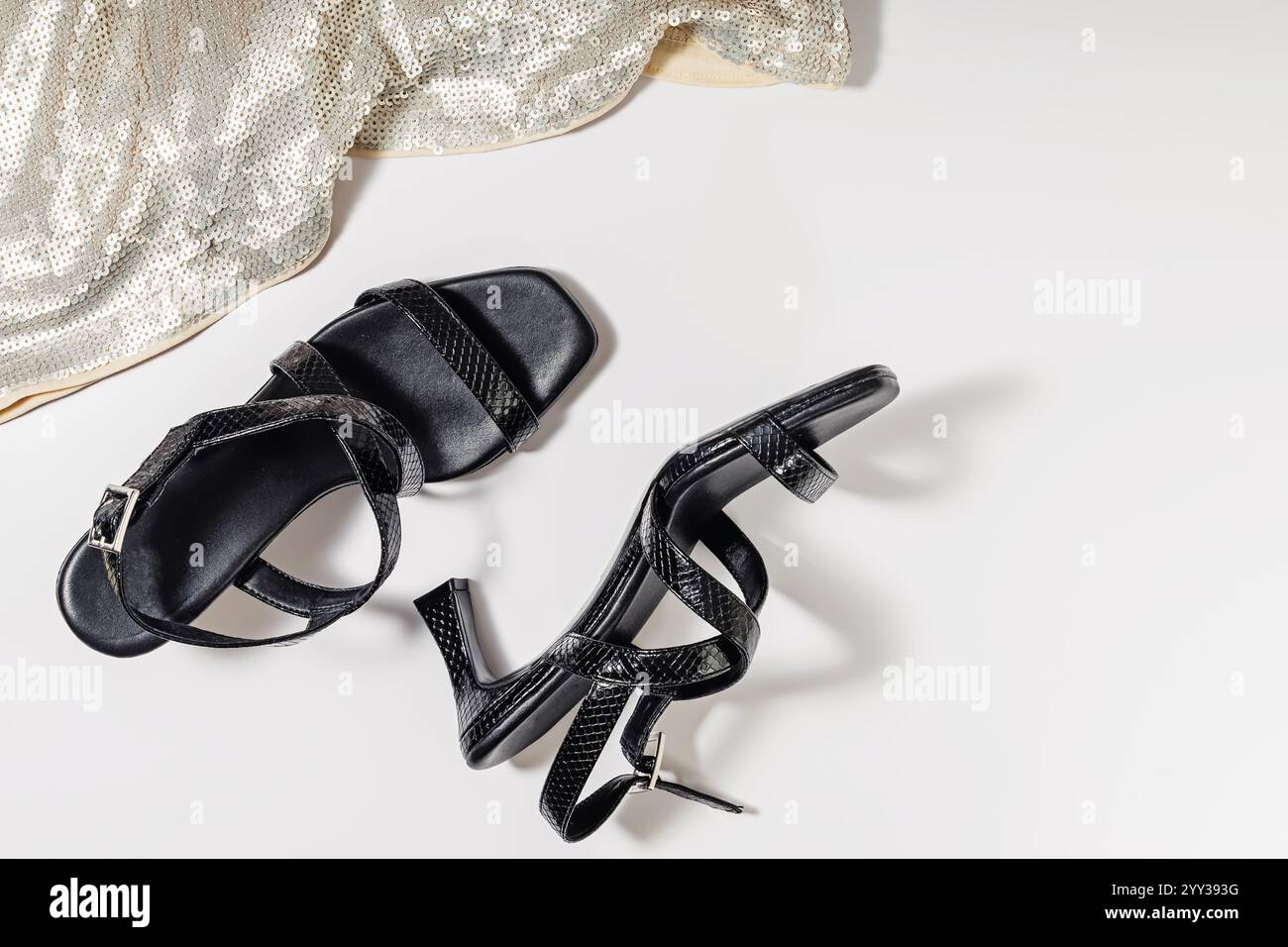 Élégantes sandales à brides en cuir noir placées sur une surface neutre avec un tissu chatoyant Banque D'Images