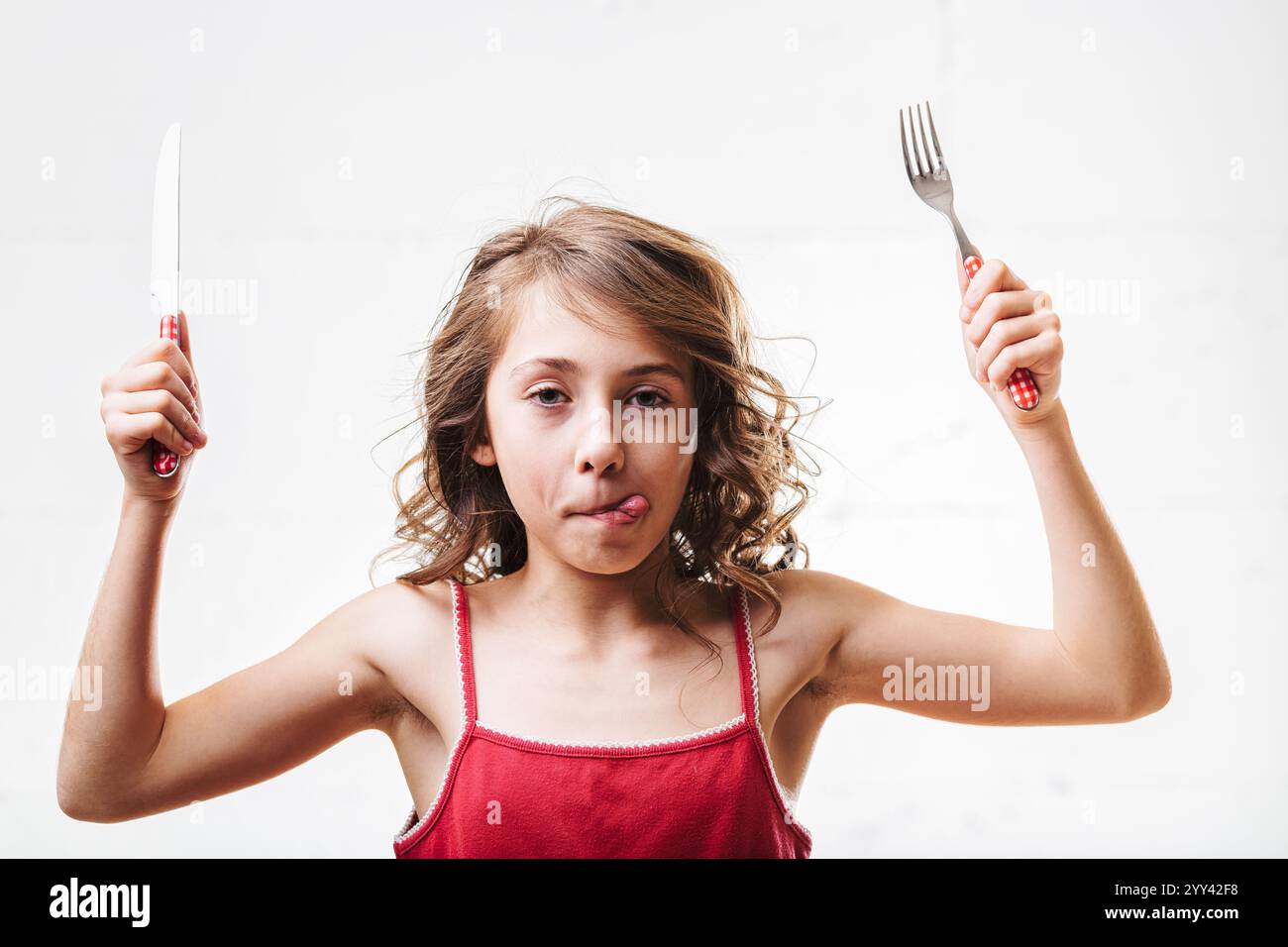 Jeune fille tenant énergiquement une fourchette et un couteau, sortant ludique sa langue, rayonne d'excitation et de joie dans un cadre animé de repas Banque D'Images