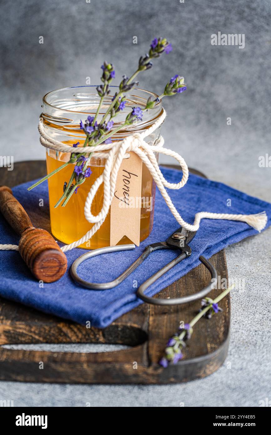 Un pot en verre de miel doré orné de fleurs de lavande et attaché avec une corde se trouve sur une planche de bois rustique Une trempette de miel en bois et ciseaux peuvent accueillir Banque D'Images