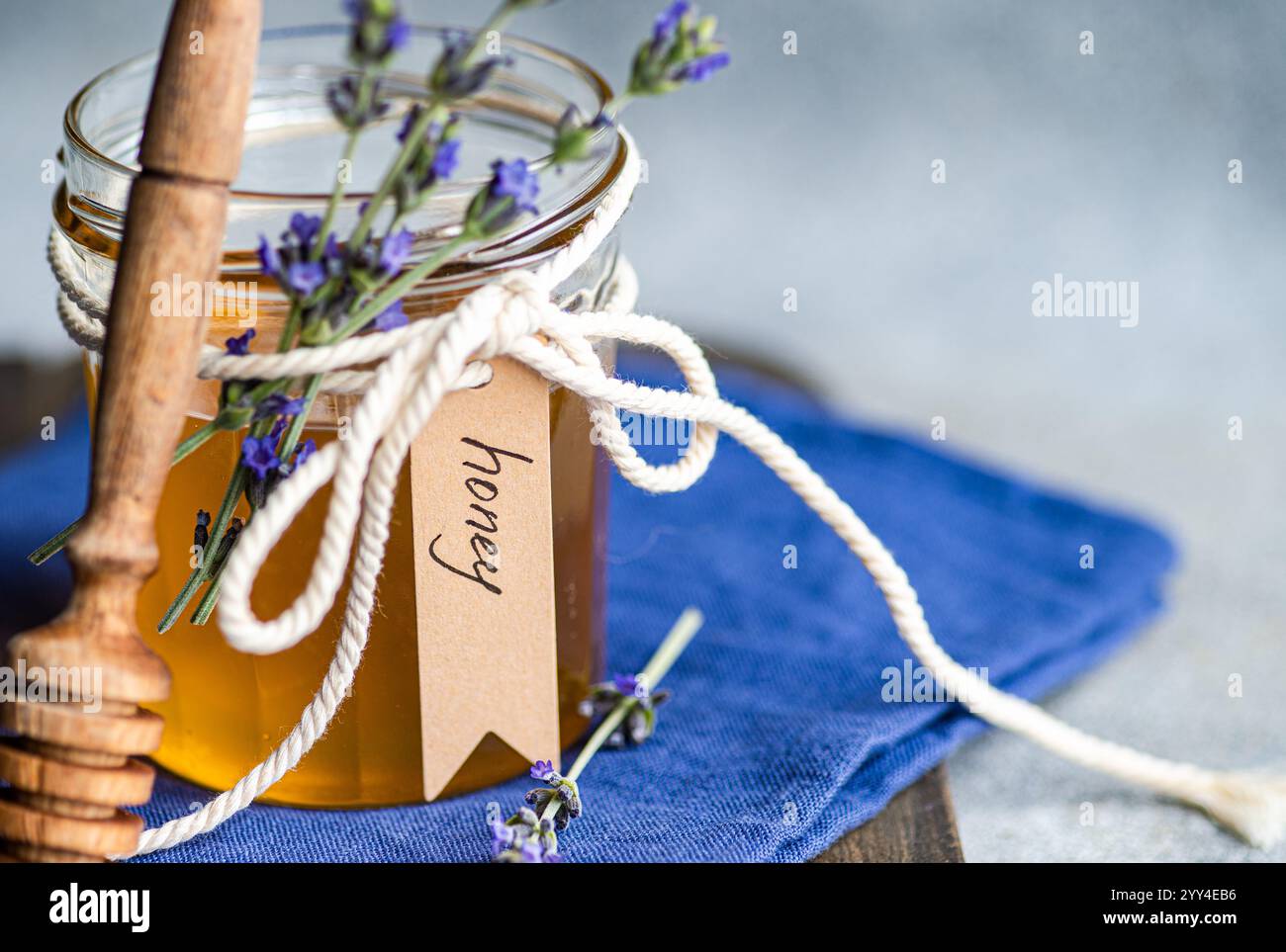 Un pot en verre de miel doré orné d'un noeud de ficelle, de petits brins de lavande, et une étiquette manuscrite Une benne de miel en bois repose à côté de lui sur un tissu bleu Banque D'Images