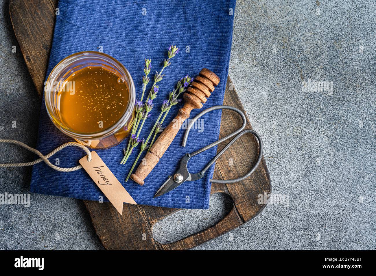 Un pot de miel doré, des brins de lavande, et une benne en bois reposent sur un tissu bleu, créant une scène culinaire sereine la planche à découper rustique et le scisso Banque D'Images
