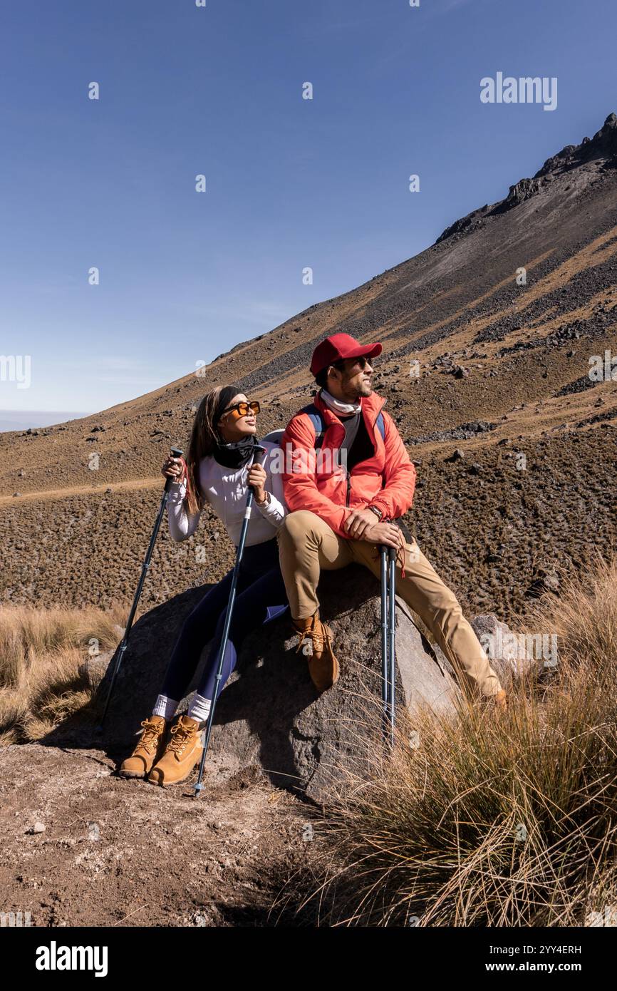 Une paire de randonneurs prenant une pause sur un sentier montagneux, ils sont assis sur un rocher tenant des bâtons de trekking, profitant de la vue panoramique sous un ciel clair Banque D'Images