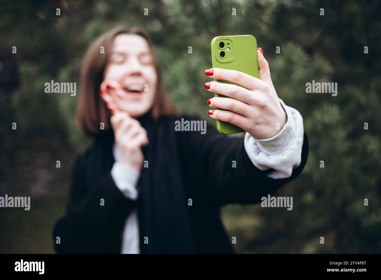 Une personne joyeuse prend un selfie avec une canne à bonbons à l'extérieur, incarnant l'esprit festif de l'étui vert du téléphone contraste avec les ongles rouges, ajoutant à la Banque D'Images