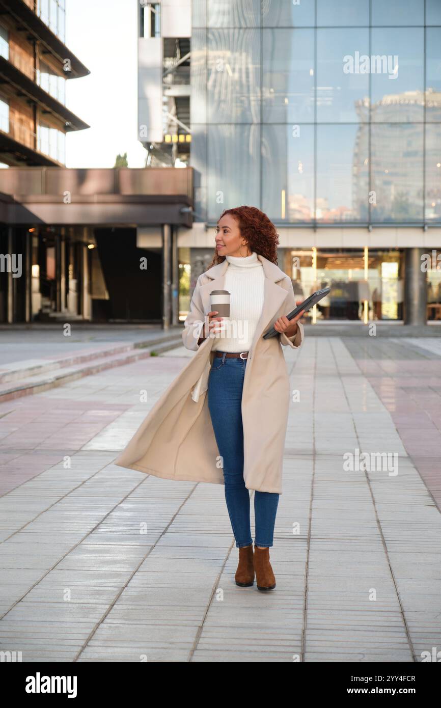 Une femme latine aux cheveux bouclés et aux traits de race mixte se promène en toute confiance dans un paysage urbain moderne. Elle porte un manteau élégant, portant un café et une note Banque D'Images
