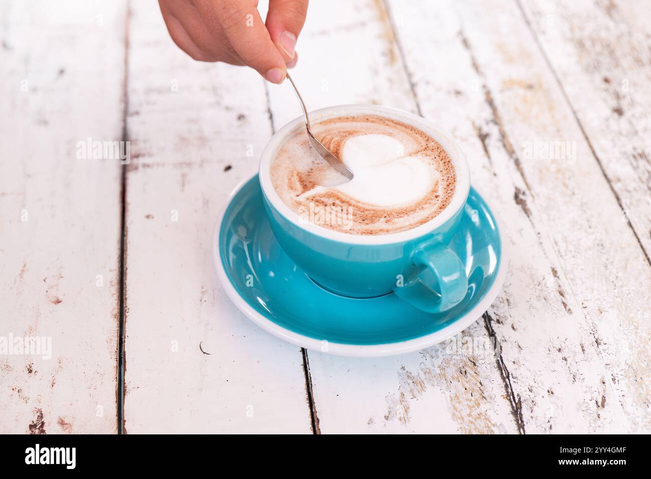 Gros plan d'une main remuant un cappuccino accompagné d'un délicat latte art en forme de cœur dans une tasse bleue. Le cadre est une table en bois rustique, mettant en évidence un co Banque D'Images
