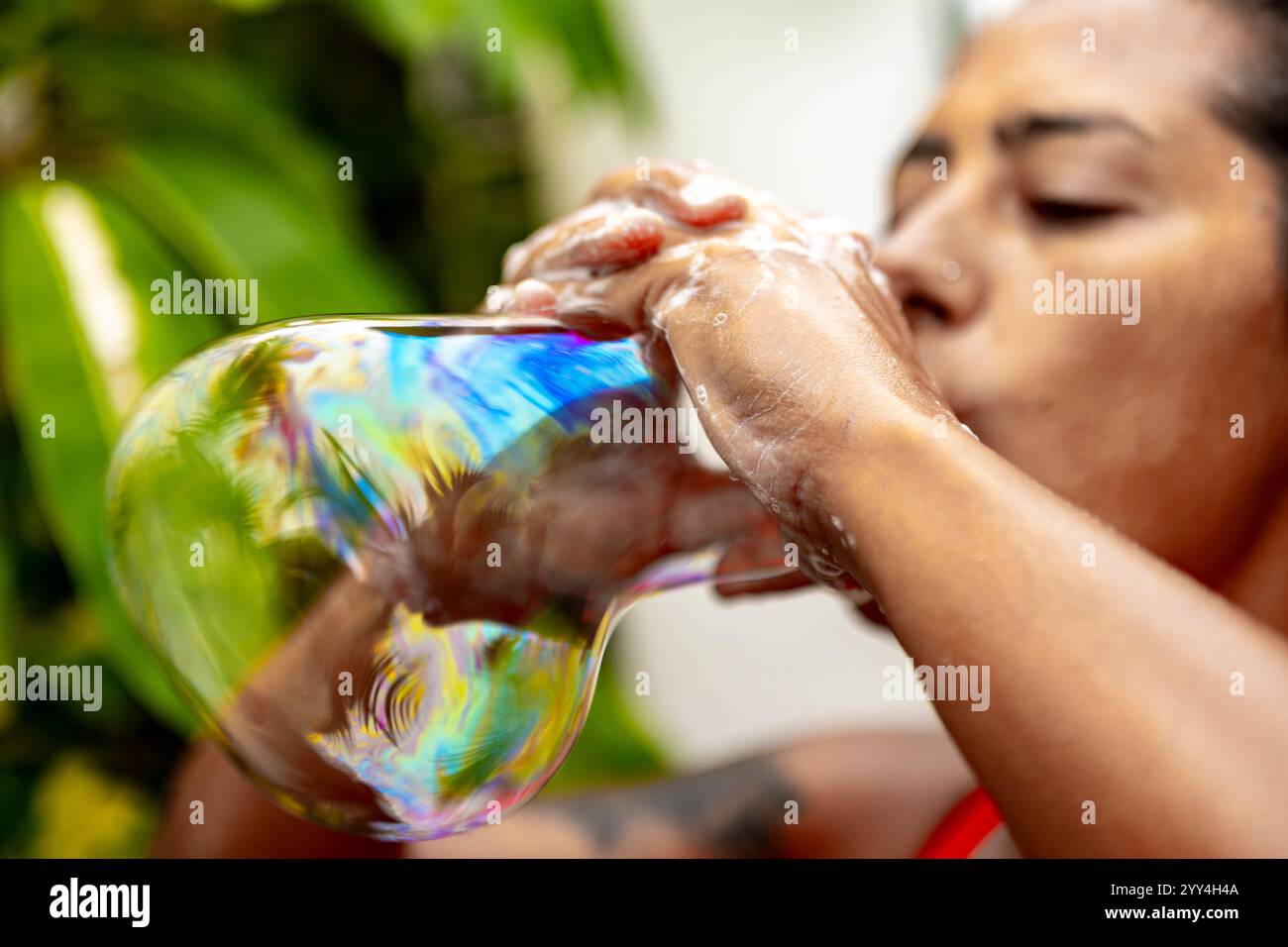 Une personne soufflant une grosse bulle de savon irisée. Les couleurs vives et les reflets capturent un moment de créativité ludique entouré de fol vert luxuriant Banque D'Images