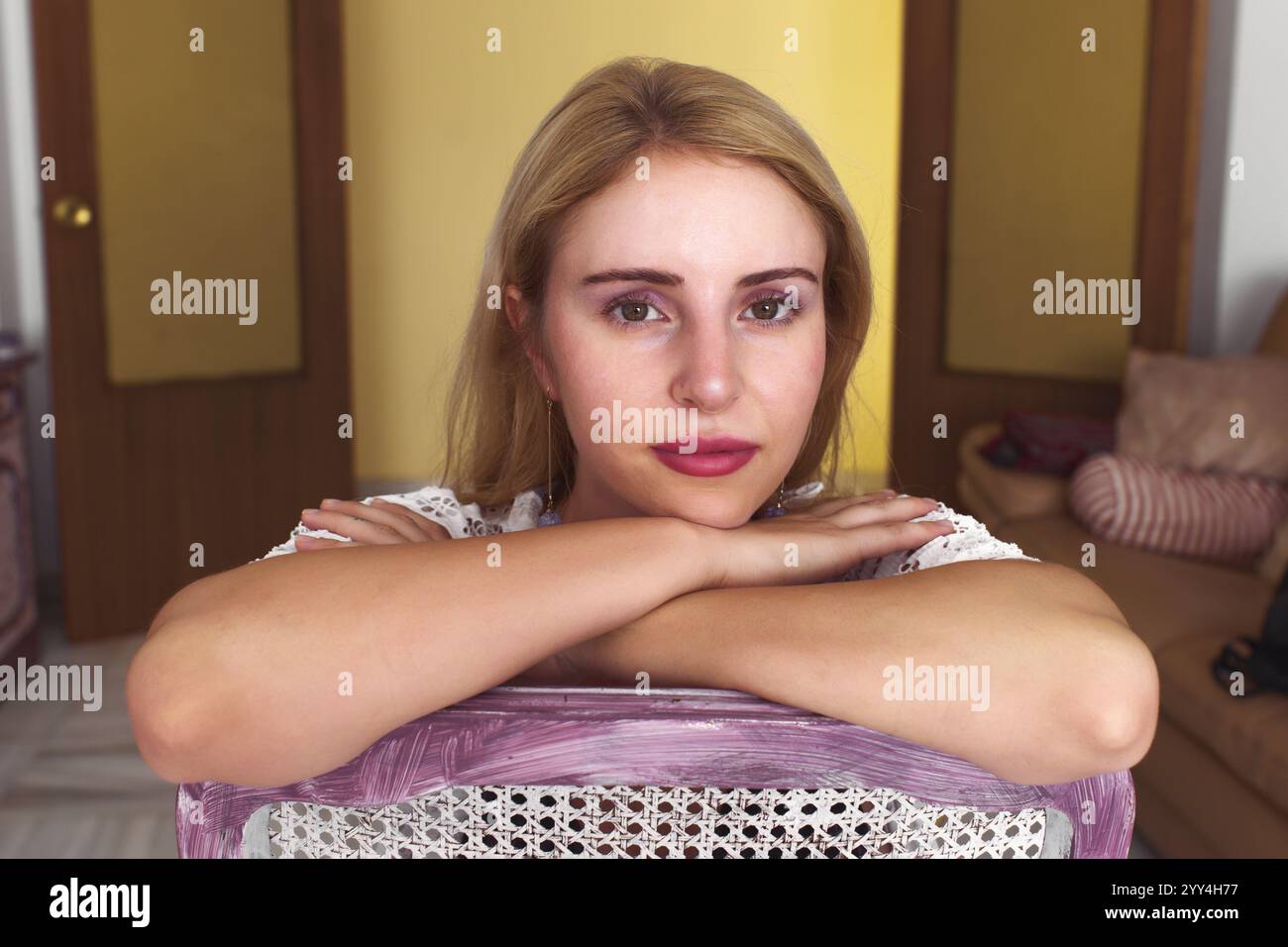 Une femme contemplative aux cheveux blonds repose ses bras sur une chaise, regardant directement vers l'avant. L'arrière-plan intérieur chaleureux et la pose détendue créent une série Banque D'Images
