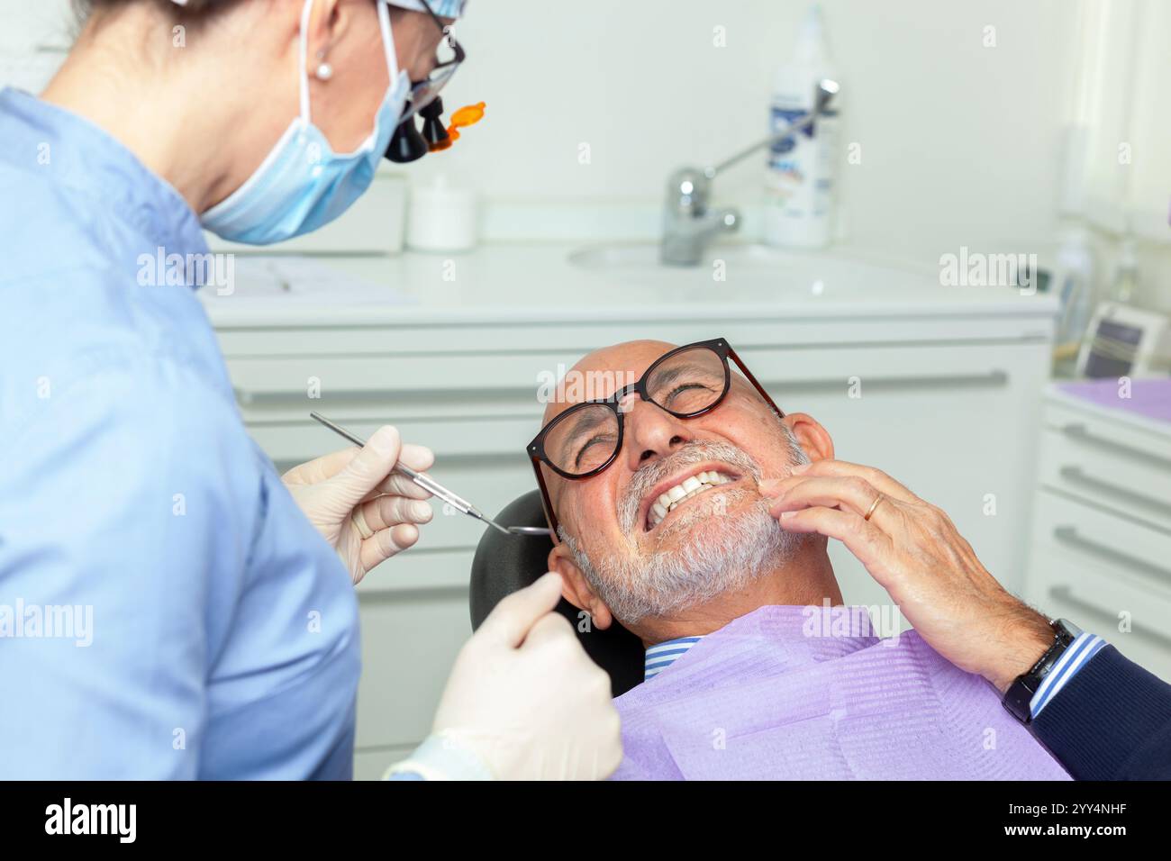 Dentiste portant une loupe et un masque chirurgical examinant un patient âgé souffrant de maux de dents dans une clinique dentaire brillante Banque D'Images