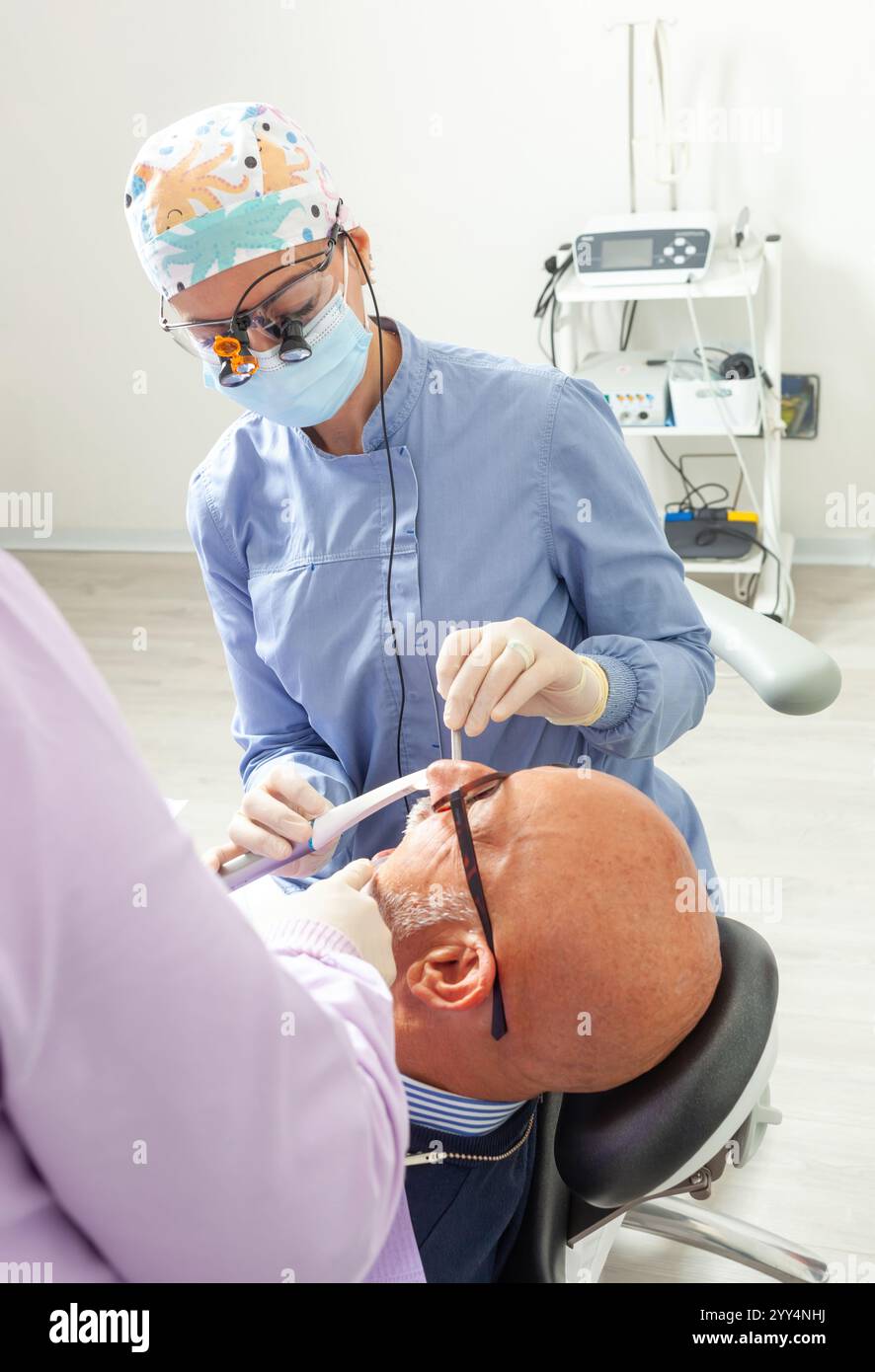Dentiste portant une loupe et un masque chirurgical examinant un patient senior en clinique dentaire Banque D'Images