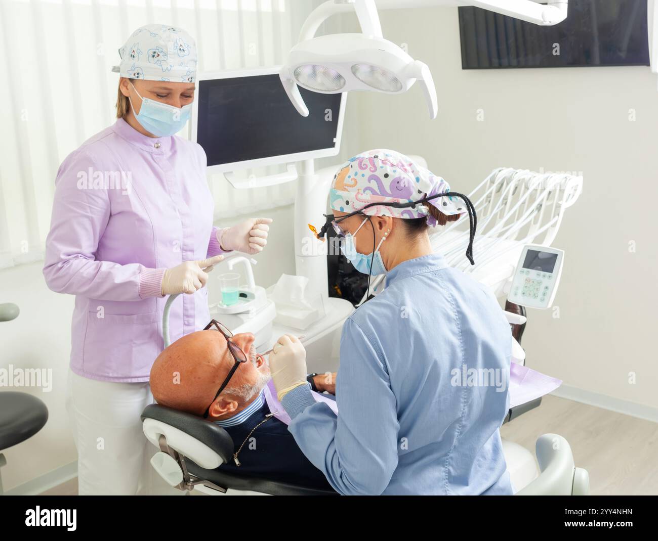 Dentiste portant des loupes effectuant un examen dentaire sur un patient masculin senior alors qu'infirmière l'assistant dans une clinique dentaire moderne Banque D'Images