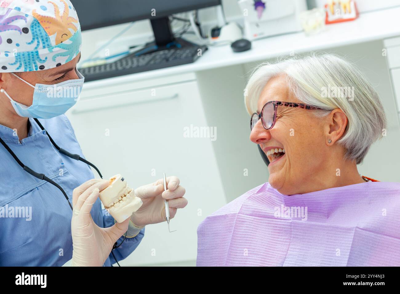 Dentiste montrant un modèle de prothèse à une femme âgée joyeuse lors d'une consultation en clinique dentaire, expliquant le traitement Banque D'Images