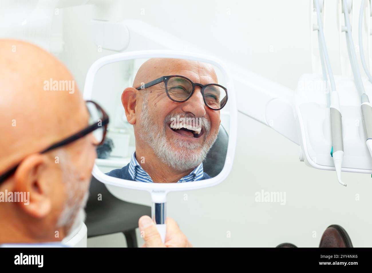 Patient senior heureux admirant son sourire parfait après un traitement d'implant dentaire, tenant un miroir dans sa main à la clinique dentaire Banque D'Images