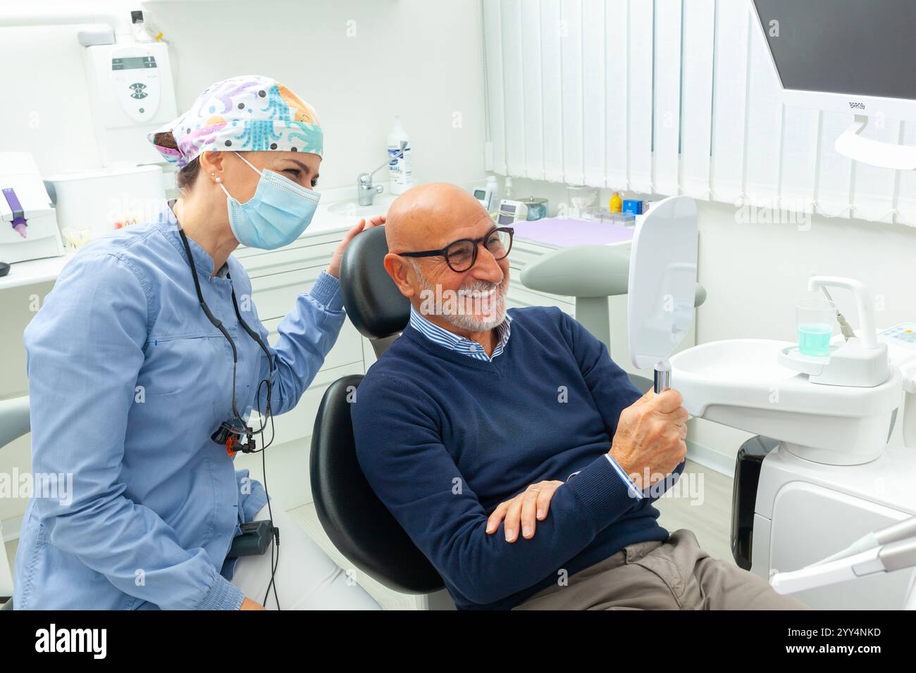 Homme âgé souriant regardant son nouveau sourire dans le miroir à la clinique dentaire, assisté par une dentiste gentille Banque D'Images