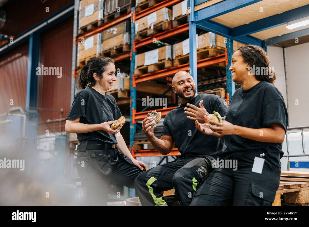 Heureux hommes et femmes ouvriers d'entrepôt mangeant des hamburgers tout en appréciant et en parlant les uns avec les autres dans l'usine Banque D'Images