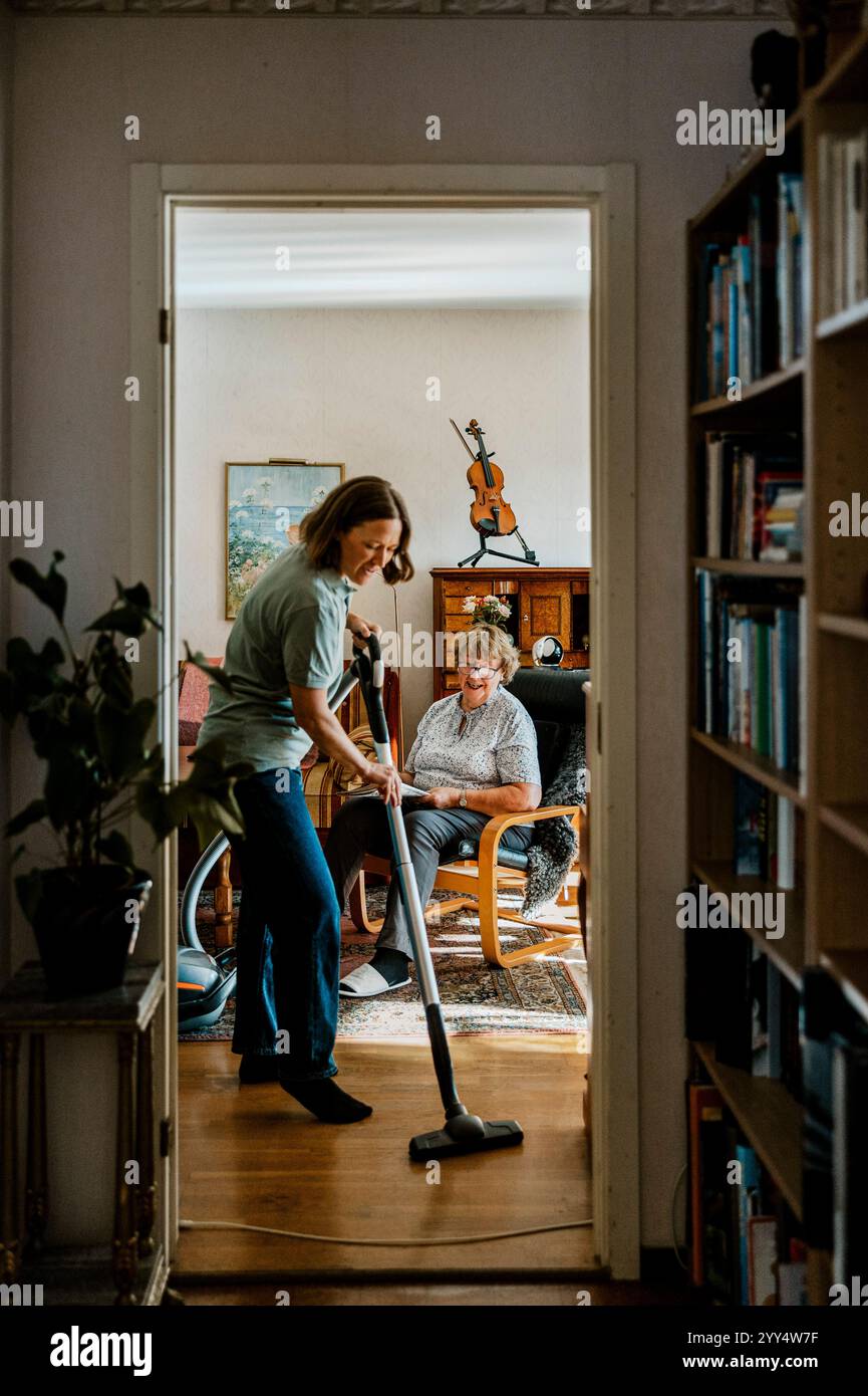 Femme âgée assise dans le salon pendant que la femme soignante passe l'aspirateur au sol à la maison Banque D'Images